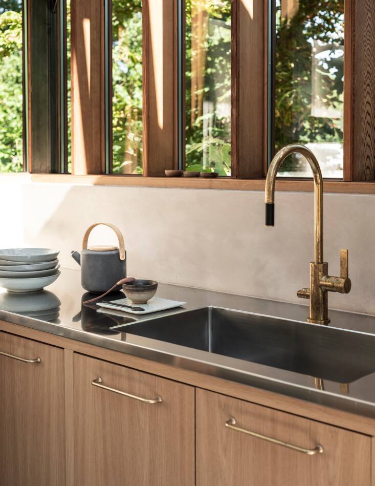 Light oak cabinets, antique brass hardware by Corston, and a brass faucet by Tapwell are a warm counterpoint to the stainless steel countertops and appliances.